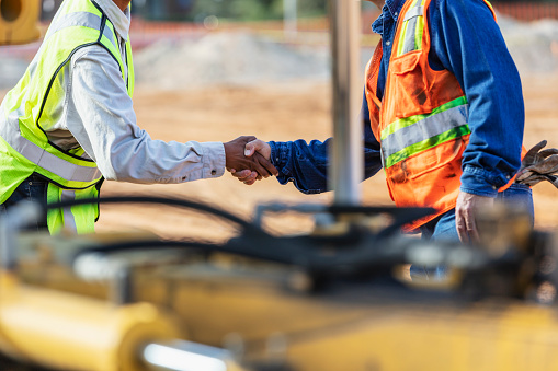 Cropped view of two multiracial construction workers shaking hands. The one in the yellow reflective vest is a mature African-American woman, in her 40s. The other one is an Hispanic man in his 50s.