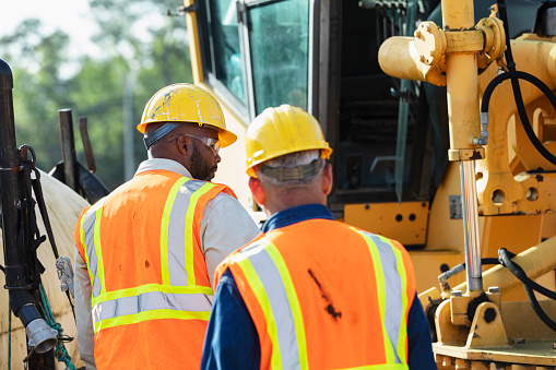 Rear view of two multiracial construction workers working at a job site. They are walking toward heavy machinery, a grader used to grade and level the ground.