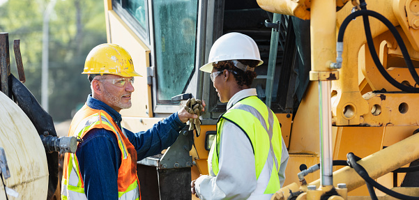 Two multiracial construction workers working at a job site. They are standing by heavy machinery, a grader used to grade and level the ground. The one in the yellow safety vest and white hardhat is a mature African-American woman in her 40s. Her coworker is an Hispanic man in his 50s.