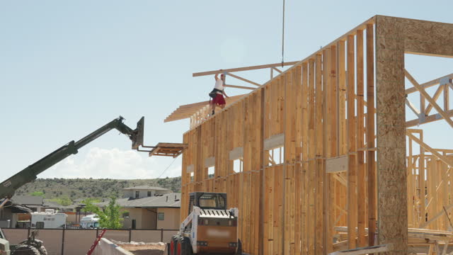 On a sunny day, a framing carpenter walks atop walls, skillfully setting and nailing down roof trusses on a residential home, ensuring a secure and durable structure is being built.