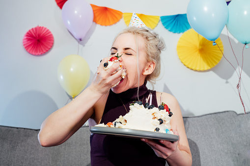 Woman in festive dress and party cap eating birthday cake with her hands, her face stained cream. A woman stuffing her face with cake. Crazy celebrating birthday party at home. Selective focus