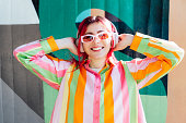 Emotional stylish young woman with pink hair and sun glasses in multicolor strippled shirt wears wireless headphones, listening to music and singing on wall background. Fashionable hipster lifestyle.