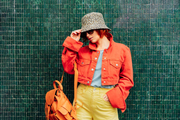 Hipster young woman in bright clothes, sun glasses, backpack bag and bucket hat posing on the green tile wall background. Urban city street fashion. Fashion blogger. Selective focus. Copy space, Hipster young woman in bright clothes, sun glasses, backpack bag and bucket hat posing on the green tile wall background. Urban city street fashion. Fashion blogger. Selective focus. Copy space bucket hat stock pictures, royalty-free photos & images