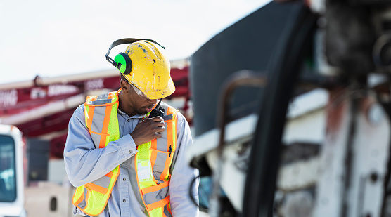 An African-American construction worker with a hard hat, ear protectors, safety glasses and reflective vest, talking on a walkie-talkie. He is a mid adult man in his 30s. This is part of a series of images and video showing workers operating concrete pump trucks at a construction site.