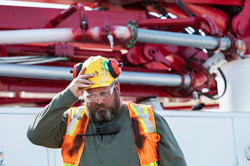 A construction worker standing in front of a concrete pump truck wearing a hard hat, protective eyewear and reflective vest, with his hand on his helmet, a serious expression. He is a mature, heavyset man in his 40s, with a beard. This is part of a series showing workers operating concrete pump trucks at a construction site.