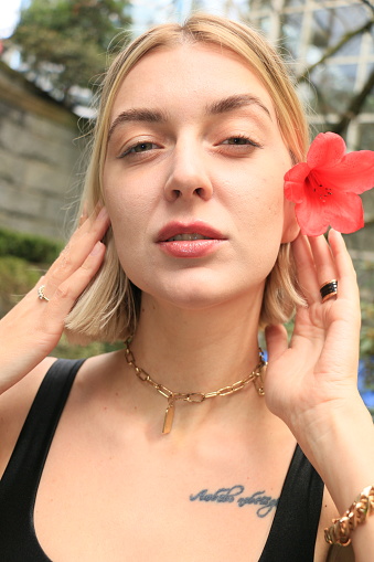 A  closeup of a Ukrainian woman with a red flower behind her ear. She is wearing makeup,, short, straight, blond hair, a necklace, rings and a tattoo.