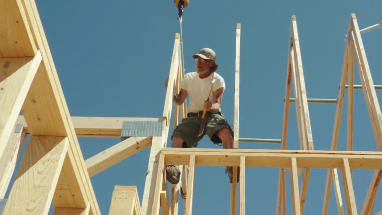 Under the bright sun, a framing carpenter walks atop the walls of a residential home, skillfully setting and nailing down roof trusses, ensuring a secure and durable structure.