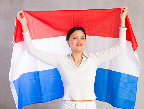 Portrait of young smiling woman in casual clothes posing with flag of Netherlands in studio
