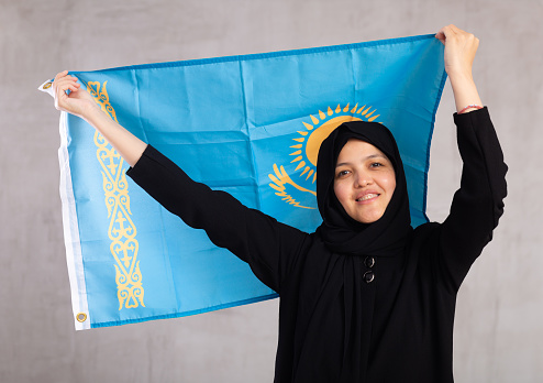 Portrait of young smiling woman in hijab posing with large flag of Kazakhstan in studio