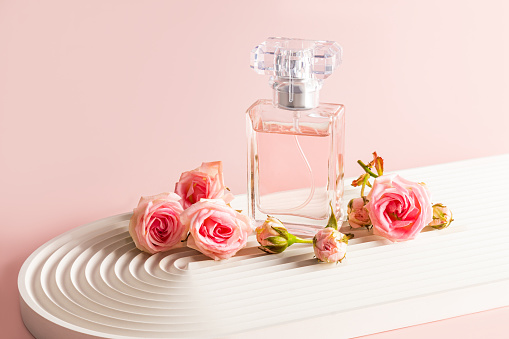 A stylish bottle of perfume stands on a white podium in the form of an arch and small pink roses. The modern image of perfume, fragrance