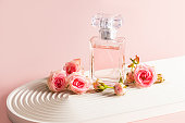A stylish bottle of perfume stands on a white podium in the form of an arch and small pink roses. The modern image of perfume, fragrance.