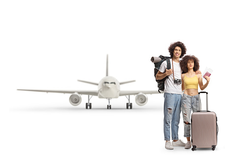 Young male and female travelers with a backpack and suitcase holding a passport in front of an airplane isolated on white background