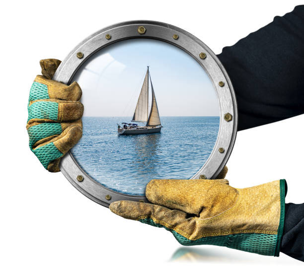 Two hands with protective work gloves hold a metal porthole with a white sailing boat in motion in the blue Mediterranean sea, Liguria, Italy, Europe.