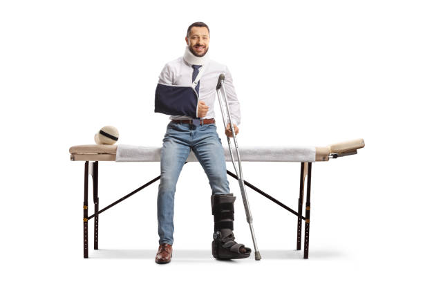 young injured man with cervical collar, orthopedic boot and arm sling sitting on a therapy table - physical injury men orthopedic equipment isolated on white imagens e fotografias de stock