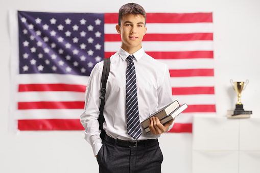 Male student in shirt and tie holding books and standing in front of a USA flag