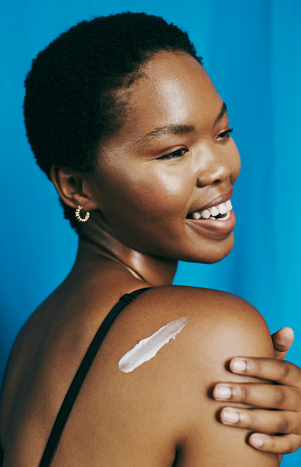 Fresh faced beauty. Happy, young black woman looking over her shoulder whilst applying body lotion. Stock photo