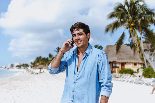 hispanic young man talking by mobile phone at the beach on vacations or holidays in Mexico Latin America, Caribbean and tropical destination