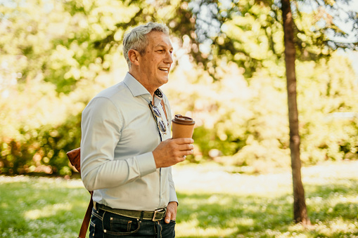 Man walking to office hanging a bag on his shoulder. Smiling businessman going to the office and drinking a takeaway coffee.