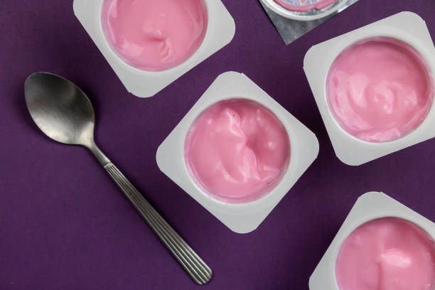 yogur rosa con sabor a frutas en vasos de plástico blanco sobre fondo púrpura brillante con tapa de aluminio plateado - vista superior foto de fondo de taza de yogur con enfoque selectivo - yogurt yogurt container strawberry spoon fotografías e imágenes de stock