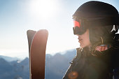 Portrait of sportswoman in helmet and mask with skis in hand, looking away, enjoying sunny frosty day, perfect day for skiing in ski resort