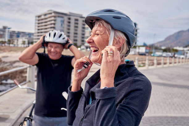 casco, risas y una pareja mayor en bicicleta al aire libre juntos para hacer ejercicio o un estilo de vida activo. verano, ejercicio o humor con un hombre y una mujer maduros riendo en el paseo marítimo durante el verano - action mature adult bicycle senior couple fotografías e imágenes de stock