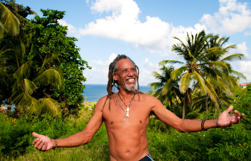 A Rastafarian man with his arms spread wide against a tropical background.