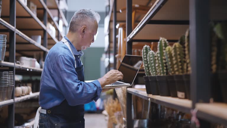 Technology has affected small businesses with entrepreneurs and their employees adapting to new systems. Japanese home decor store owner with a laptop in a warehouse for check delivery and customer orders, stock for the supply to customer need.