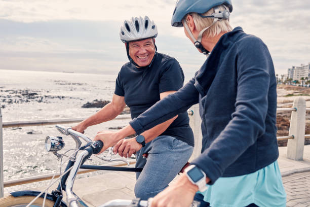 bicicletta, ciclismo e spiaggia con una coppia di anziani che pedala all'aperto sul lungomare durante l'estate per l'esercizio. bicicletta, fitness o tempo libero con un uomo e una donna maturi che fanno un giro insieme sulle loro biciclette - mature adult bicycle senior adult heterosexual couple foto e immagini stock