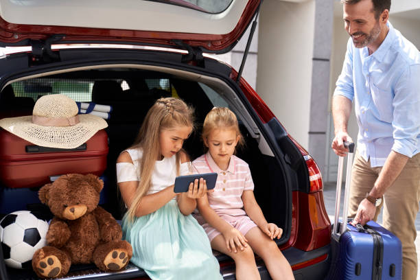 istock Two caucasian children browsing mobile phone while sitting in car trunk and father packing luggage 1491218431
