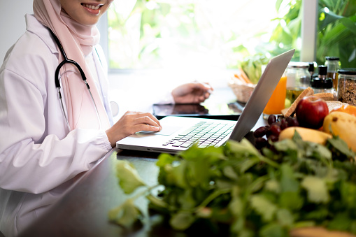 Asian Muslim nutrition consulting using laptop on table with green vegetable and fruits background beside window.