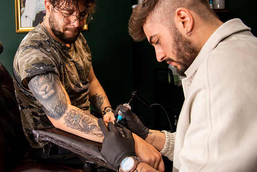 Close up of professional tattooer artist doing picture on hand of man by machine black ink from a jar. Tattoo art on body. Equipment for making tattoo art.