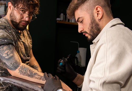Close up of professional tattooer artist doing picture on hand of man by machine black ink from a jar. Tattoo art on body. Equipment for making tattoo art.