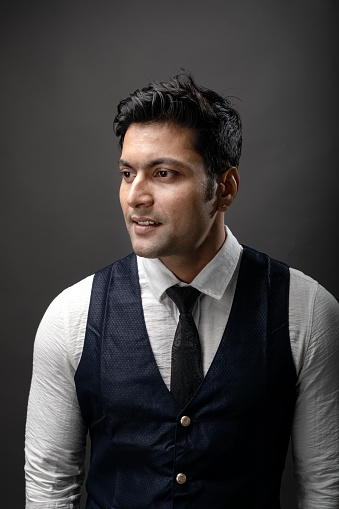 Studio shot of cheerful, young, handsome Indian male model in formal wear wearing white shirt, black tie, black coat against grey background. Male model. Fashion Portrait.