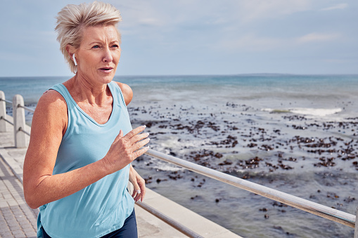 Senior woman, exercise and running at beach promenade, sky mockup or energy of health, wellness and workout. Elderly female, earphones and fitness at ocean of sports, cardio fitness or strong mindset