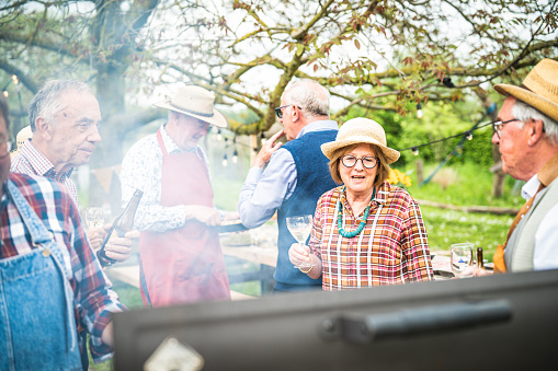 Group of different elderly people preparing barbecue and sipping wine and beer, preparing a lunch and an outdoor party in the garden, concept of healthy and happy elderly people