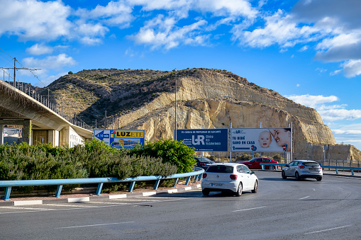 Alicante, Spain - April 30, 2023: Cars driving in a roundabout area of a highway with greenery, a bridge across the roundabout, and a mountain in the background under a cloudy blue sky. No People are on the scene.