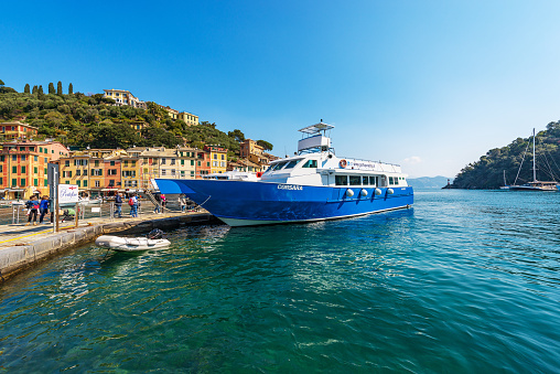 Portofino, Liguria, Italy - April 8th, 2023: Ferry with tourists in the famous village of Portofino on a sunny spring day, luxury tourist resort in Genoa Province, Liguria, Italy, Europe. Port and colorful houses, Mediterranean sea (Ligurian sea). The village of Portofino is a famous vacation resort of the Liguria for its picturesque harbor and colorful houses that are reflected in the sea. In ancient times it was a small fishing village. Many celebrity and artists frequent this Italian seaside resort.