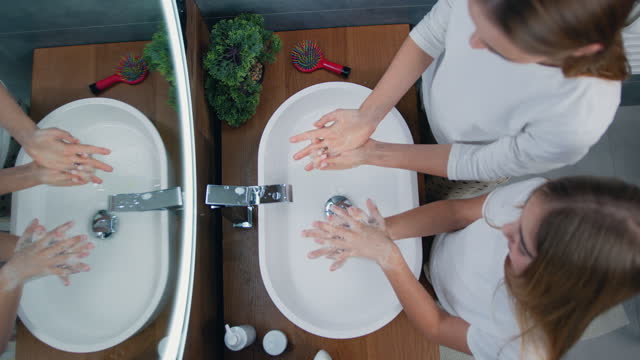Top view of two beautiful sisters washing and soaping their hands with soap in modern bathroom at home. Liquid antibacterial soap and foam. Infection prevention.