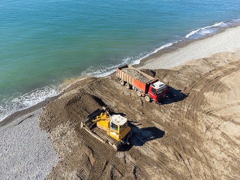 Drone view of a working bulldozer and a truck with small stones on the seashore on a sunny day. Concept of strengthening the embankment, beach of the sea.