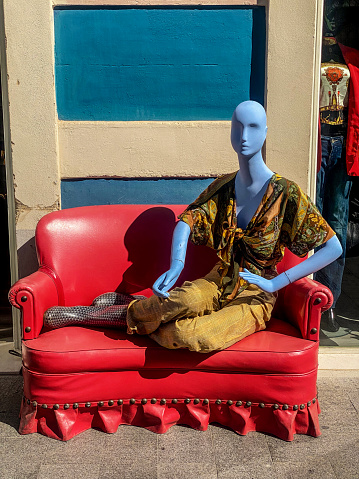 Valencia, Spain - March 4, 2023: Stylish blue mannequin reclined over red couch in front of vintage clothing store. This shops offer fashionable garments for people that like dressing for a reasonable price.