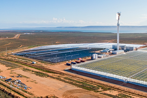 Port Augusta, Australia - September 23, 2022: Aerial view Sundrop Concentrated Solar Power (CSP) plant (or concentrated solar thermal) with associated 20 hectare greenhouse and scenic Spencer Gulf backdrop.  The receiver is glowing brightly from the reflected sunlight from the 23,000 mirrors. The system uses the sun's energy to produce freshwater from seawater (desalination) for irrigation and also produces electricity to heat and cool the crops in the greenhouse. The Sundrop 20 hectare sustainable greenhouse, produces fresh truss tomatoes for a major Australian supermarket.