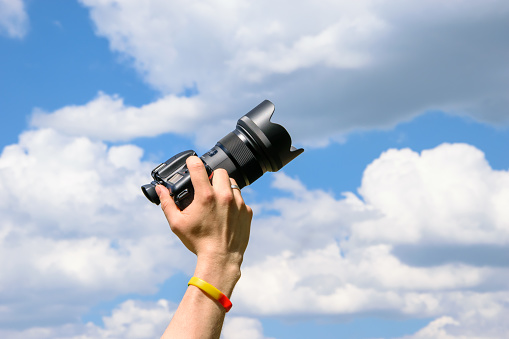 Hand holding a photo camera on a blue sky background.  Clouds coming out of the camera.  artistic shot from below