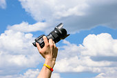 Hand holding a photo camera on a blue sky background.  Clouds coming out of the camera
