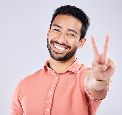 Portrait, smile and peace sign of Asian man in studio isolated on a gray background. Face, v hand emoji and happy, smiling and excited, young and confident male model with gesture or peaceful symbol.