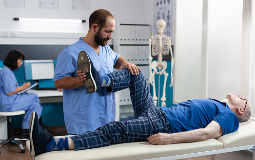 Adult with knee injury receiving osteopathic assistance for physical recovery. Assistant massaging senior patient with leg muscle pain, helping to relieve pressure for kinetotherapy.