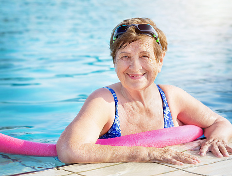 Active senior (elderly) woman (over age of 50) in sport goggles, swimsuit and with swim noodles in swimming pool before water aerobics smiling in summer day. Healthy lifestyle.