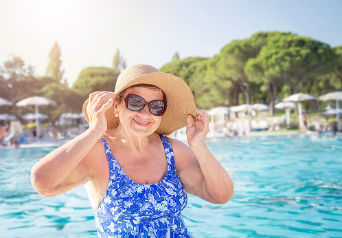 Senior (elderly) woman (over age of 50) in swimsuit, hat, sunglasses relaxing in resort swimming pool. Happy retired female enjoying summer vacation in hotel. All-inclusive.