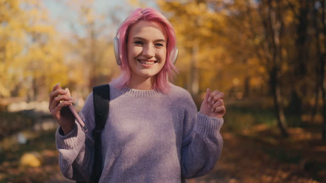 Cheerful pink-haired woman in headphones walks in fall park, enjoying music, app