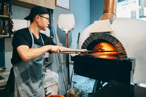 A male Asian restaurant chef prepares for the opening by igniting the fire in the wood-fired pizza oven.He uses a fire poker to push and maintain the wood in the oven, keeping the flames alive.
