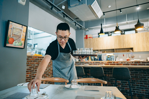 One Asian male pizza shop owner carefully places utensils on the table before today's business hours, creating a comfortable dining environment for the customers. He prepares to serve and ensures that they have an enjoyable dining experience.
